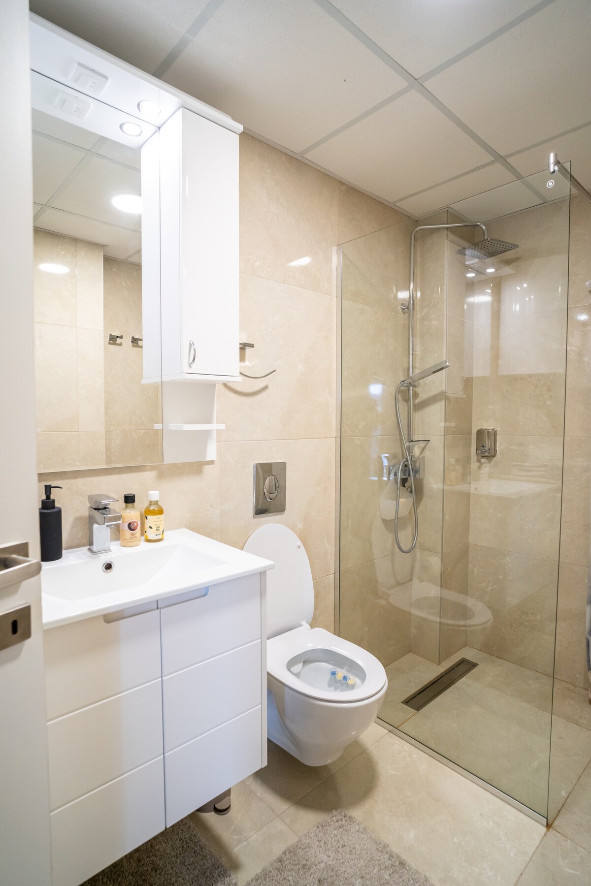A modern bathroom features light taupe tiles and a glass shower enclosure. A white vanity with several drawers is positioned beside a toilet. Shelves hold various toiletries, while overhead lights provide ample illumination.