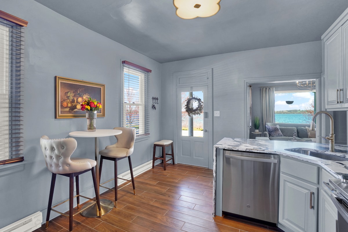 The kitchen features light blue walls with a warm wood floor. A small round table is surrounded by two upholstered chairs. Natural light fills the space through large windows, highlighting the modern cabinetry and stainless steel appliances.