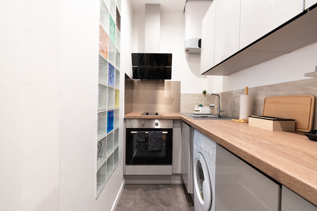 A modern kitchen is displayed with sleek cabinetry and a light wood countertop. An oven and stovetop are positioned adjacent to a washing machine. A colorful glass tile wall adds visual interest, while essential kitchen items are neatly arranged, ensuring a functional cooking space.