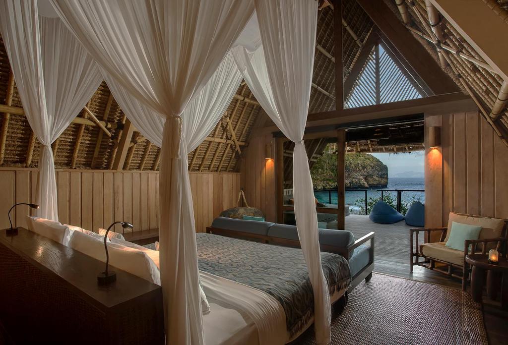 The spacious bedroom features a super-king bed with a mosquito net draping above. Light filters through the open doors, revealing a patio with ocean views. A comfortable sofa and seating area are positioned near the entrance, surrounded by natural wooden walls.