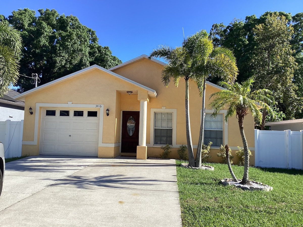 An inviting single-story home features a warm yellow exterior. A well-kept front yard includes palm trees and lush greenery, while a concrete driveway leads to a garage. Large windows allow natural light to illuminate the entrance, enhancing the welcoming appearance of the property.