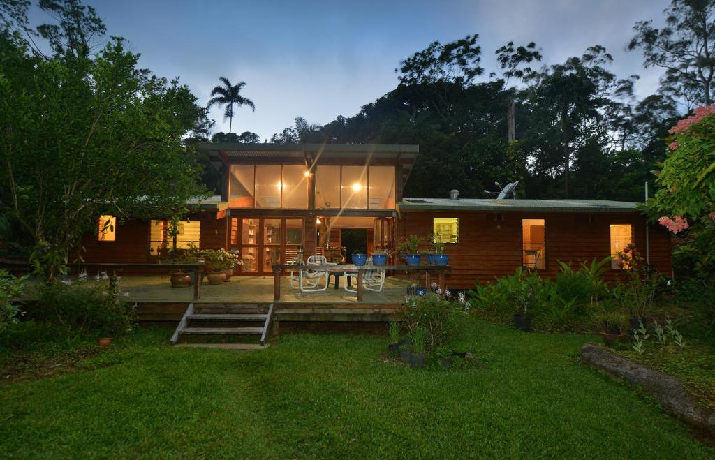 A two-story timber house is nestled among lush greenery, with large glass doors providing access to a spacious deck. Potted plants frame the entrance, and inviting outdoor seating is visible. The surrounding landscape showcases a tranquil and natural setting illuminated by soft evening light.