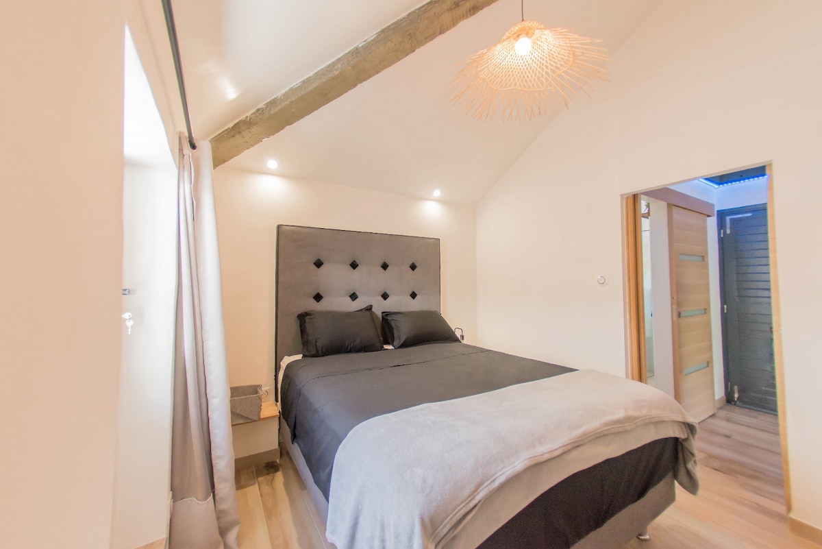 The bedroom features a spacious bed dressed in dark linens, complemented by a decorative headboard with diamond tufting. Soft lighting from built-in fixtures illuminates the room, while a textured pendant light adds a contemporary touch. A window with curtains allows for natural light.