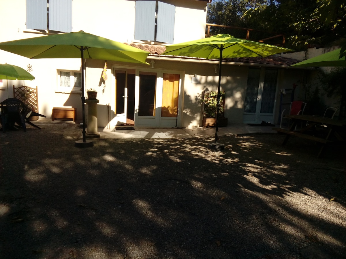 An outdoor area features a gravel surface shaded by two large green umbrellas. The entrance of the gîte is visible, flanked by windows that invite natural light. Potted plants add a touch of greenery, while patio furniture offers a space for relaxation.