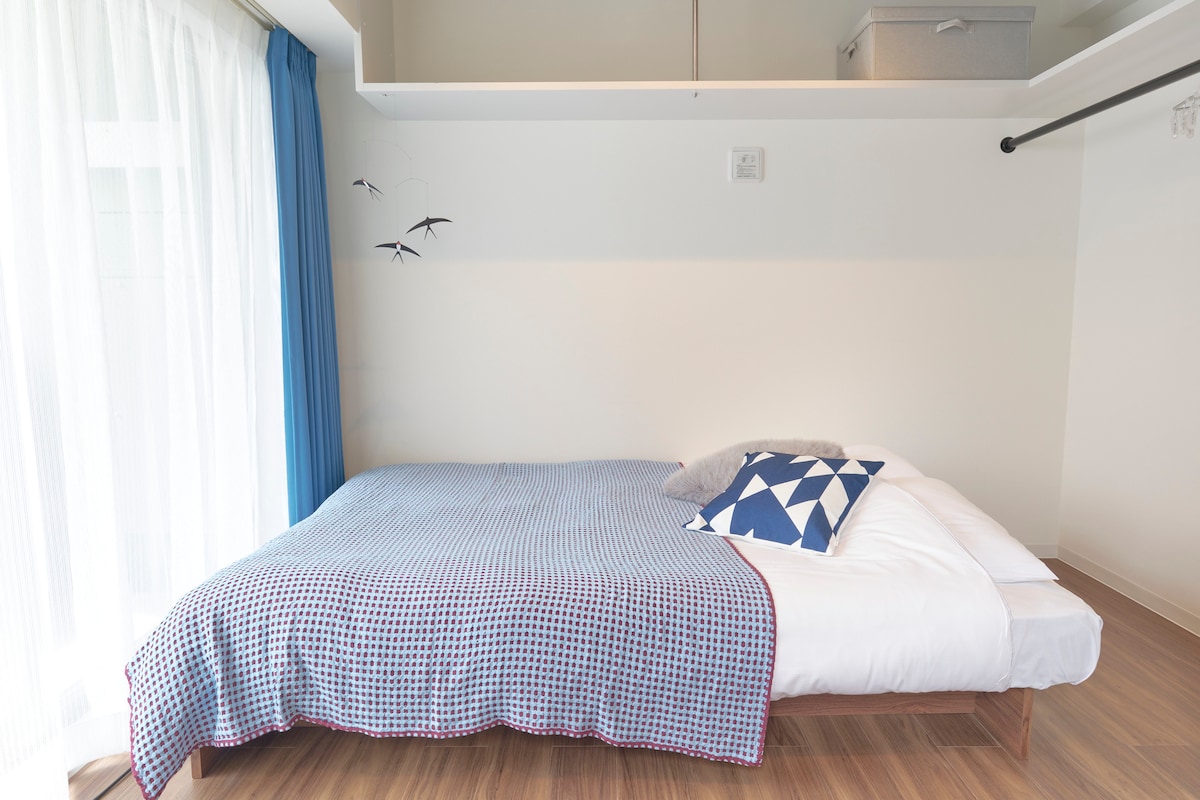 A spacious sleeping area features a large bed with a blue checkered blanket and decorative pillows. Soft light enters through sheer white curtains, and a small shelf above offers additional storage. Minimalist decor is complemented by a few wall decals of birds.
