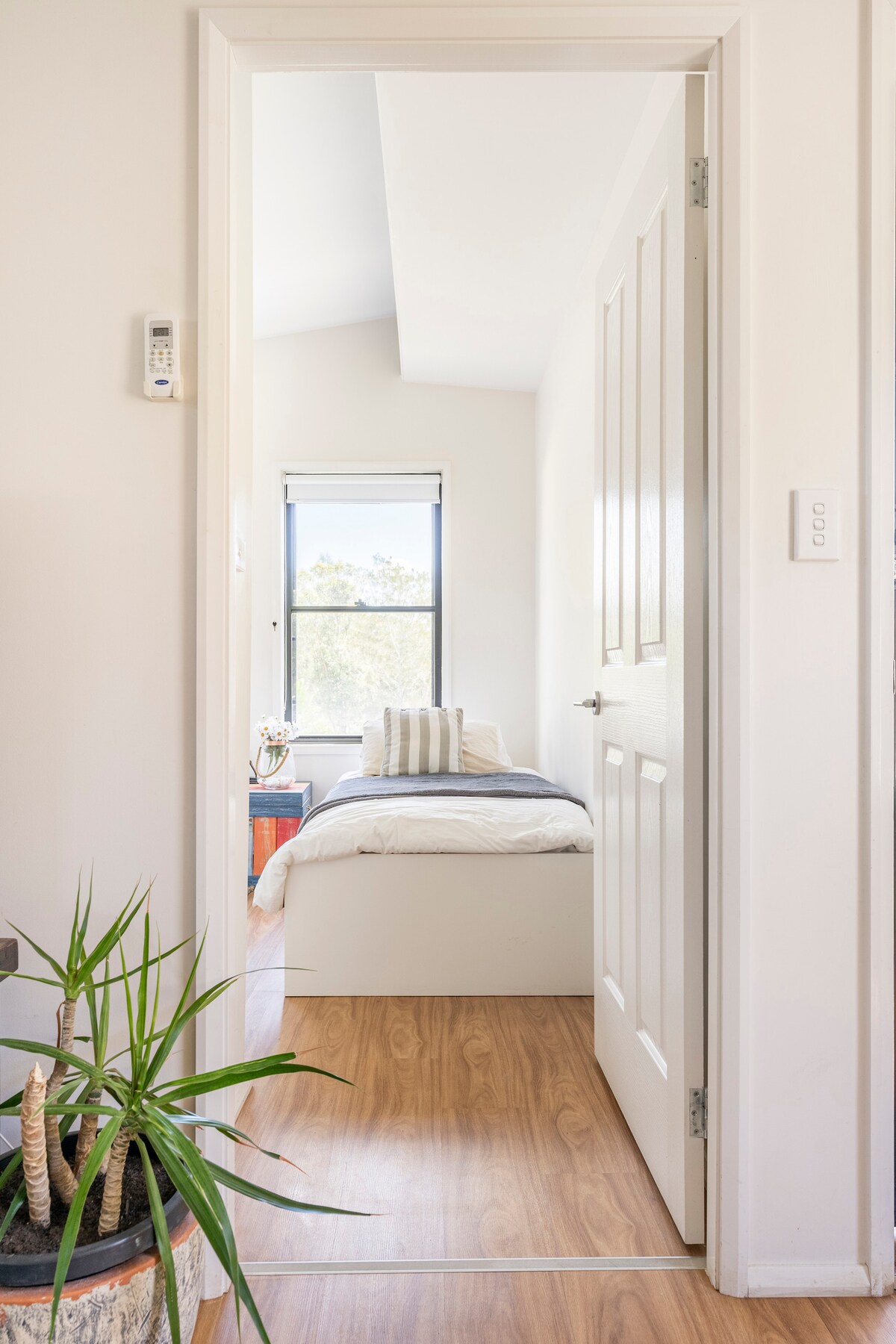 A view through an open door reveals a serene bedroom with light hardwood flooring and a bed dressed in neutral linens. A window allows natural light to illuminate the space, while a small plant is positioned strategically near the entrance.