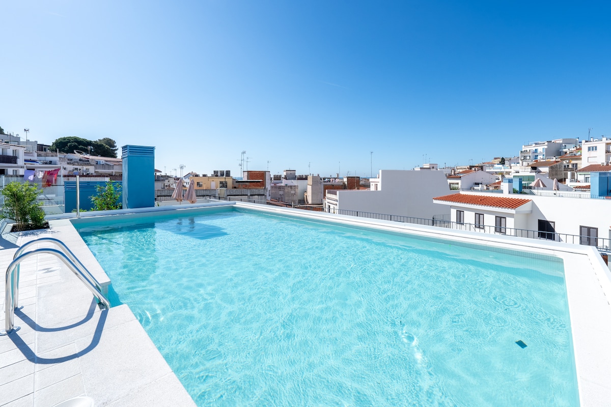 A serene rooftop swimming pool reflects the clear blue sky, framed by a modern deck and a gentle slope. Nearby, a small plant adds a touch of greenery, complementing the surrounding urban landscape with buildings in the background.