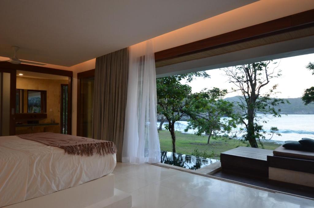 A spacious bedroom is seen with a large bed dressed in neutral tones. Floor-to-ceiling windows provide views of the sea and surrounding greenery, allowing natural light to fill the space. A comfortable seating area is situated near the window.