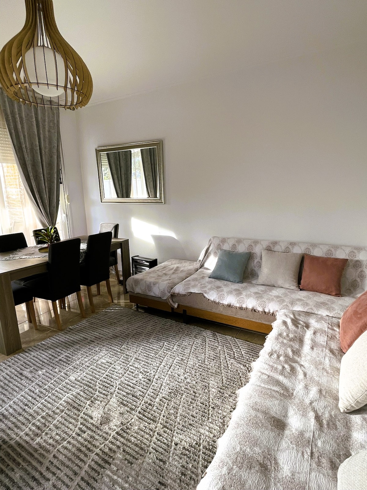 A bright living area features a light-toned sofa adorned with several decorative pillows. A wooden dining table is surrounded by dark chairs, complementing the warm tones of the space. Natural light enters through windows, reflecting off a decorative mirror on the wall.