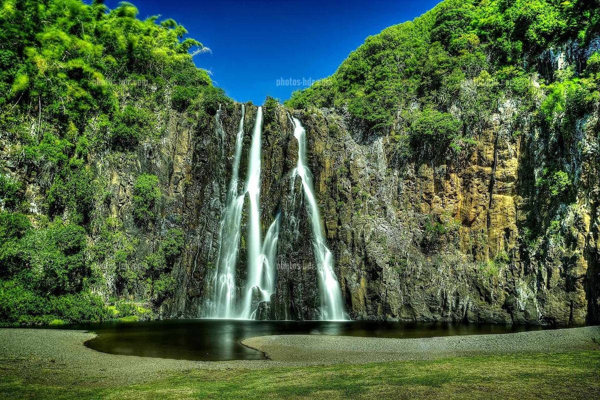 A serene waterfall cascades down a rocky cliff, creating gentle streams that flow into a calm pool below. Lush greenery surrounds the area, enhancing the natural landscape. The clear blue sky serves as a backdrop, contributing to the peaceful environment.