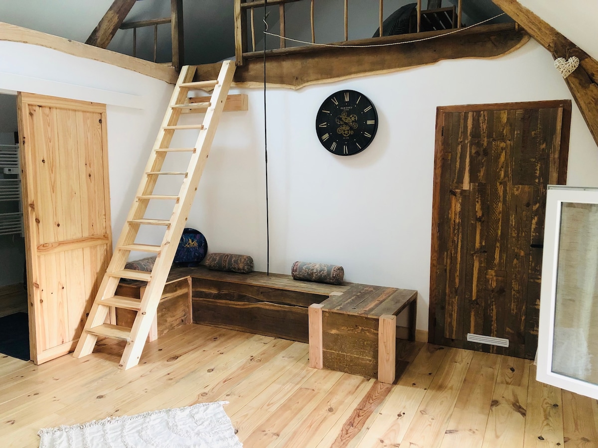A wooden ladder leads to a loft area, while a rustic wooden bench with cushions is positioned against the wall. A large wall clock is mounted above the bench, contributing to the simple and functional design of the space. Natural wood flooring creates a warm ambiance.
