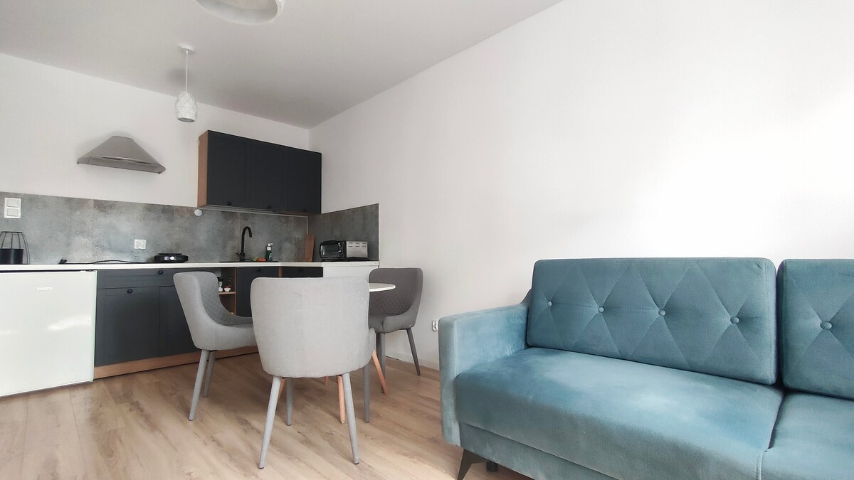 A modern kitchen space is shown, featuring dark cabinetry and a light-colored countertop. A round dining table with four grey chairs is present, complemented by a cozy blue sofa positioned nearby. Large windows allow natural light to enhance the room's bright atmosphere.