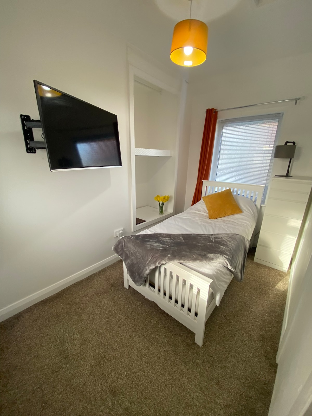 A cozy bedroom is equipped with a single bed covered in a light grey blanket. A bright orange accent pillow adds a pop of color. A wall-mounted TV is positioned across from the bed, and sunlight filters through the window, illuminating the space. A small dresser and shelves are also visible.