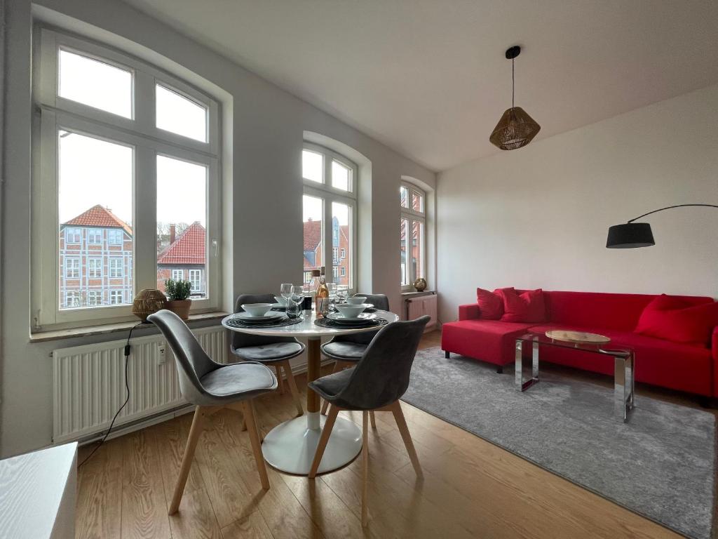 A dining area features a round table set for four, surrounded by stylish chairs. Natural light floods in through large windows, showcasing views of nearby historical buildings. A vibrant red sofa adds a pop of color, complemented by a soft gray rug beneath.