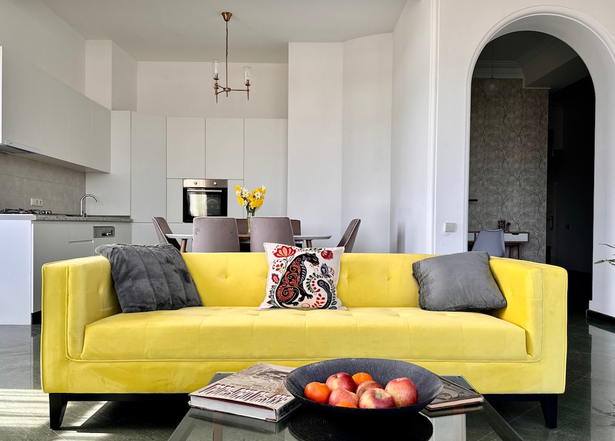 A bright yellow sofa is positioned centrally in the open-plan living area, complemented by decorative pillows. A coffee table with a bowl of apples and magazines rests in front. The modern kitchen is visible in the background, with a dining area that includes several chairs.