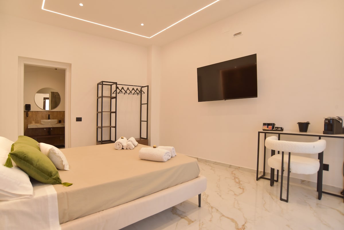 The spacious sleeping area features a large bed with neutral-toned bedding, accompanied by two neatly rolled towels. A flat-screen television is mounted on the wall. A clothing rack is positioned in the corner, and a small table with two chairs and a coffee maker is visible.