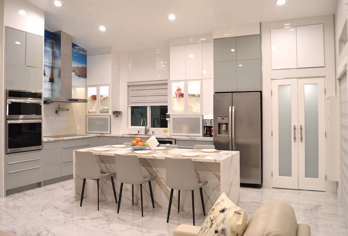 A modern kitchen features sleek cabinets and stainless-steel appliances, with ample counter space highlighted by a marble island. The dining area includes four light-colored chairs around the island, complemented by decorative fruit on a bowl. Soft lighting creates a welcoming atmosphere.
