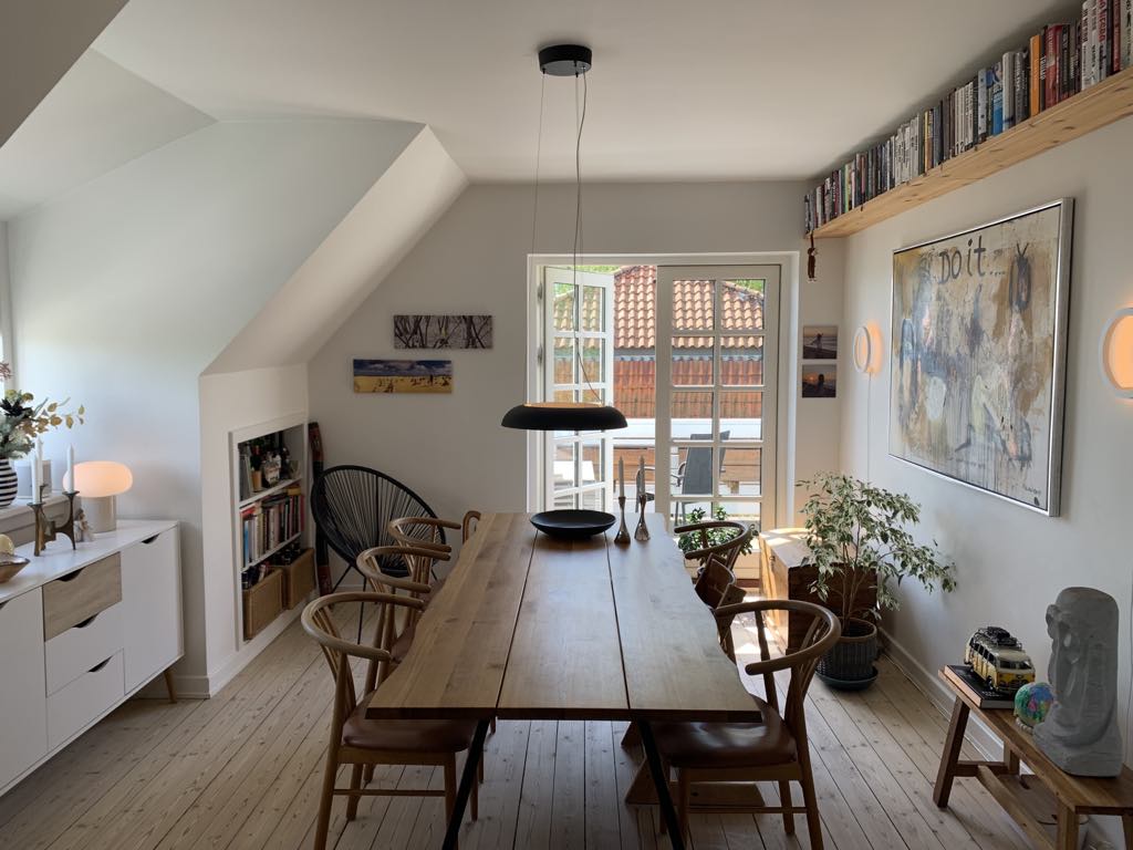 The dining area features a large wooden table surrounded by six chairs, illuminated by a pendant light. Bookcases line the walls, filled with books and decor. A potted plant adds greenery, while natural light floods the space through double doors leading to a balcony.
