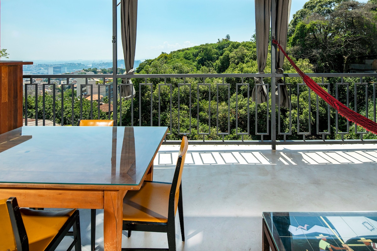 An outdoor area features a wooden dining table surrounded by chairs. A hammock is seen nearby, offering a relaxing spot. Curtains frame the space, and a lush green hillside is visible in the background, highlighting the connection with nature.