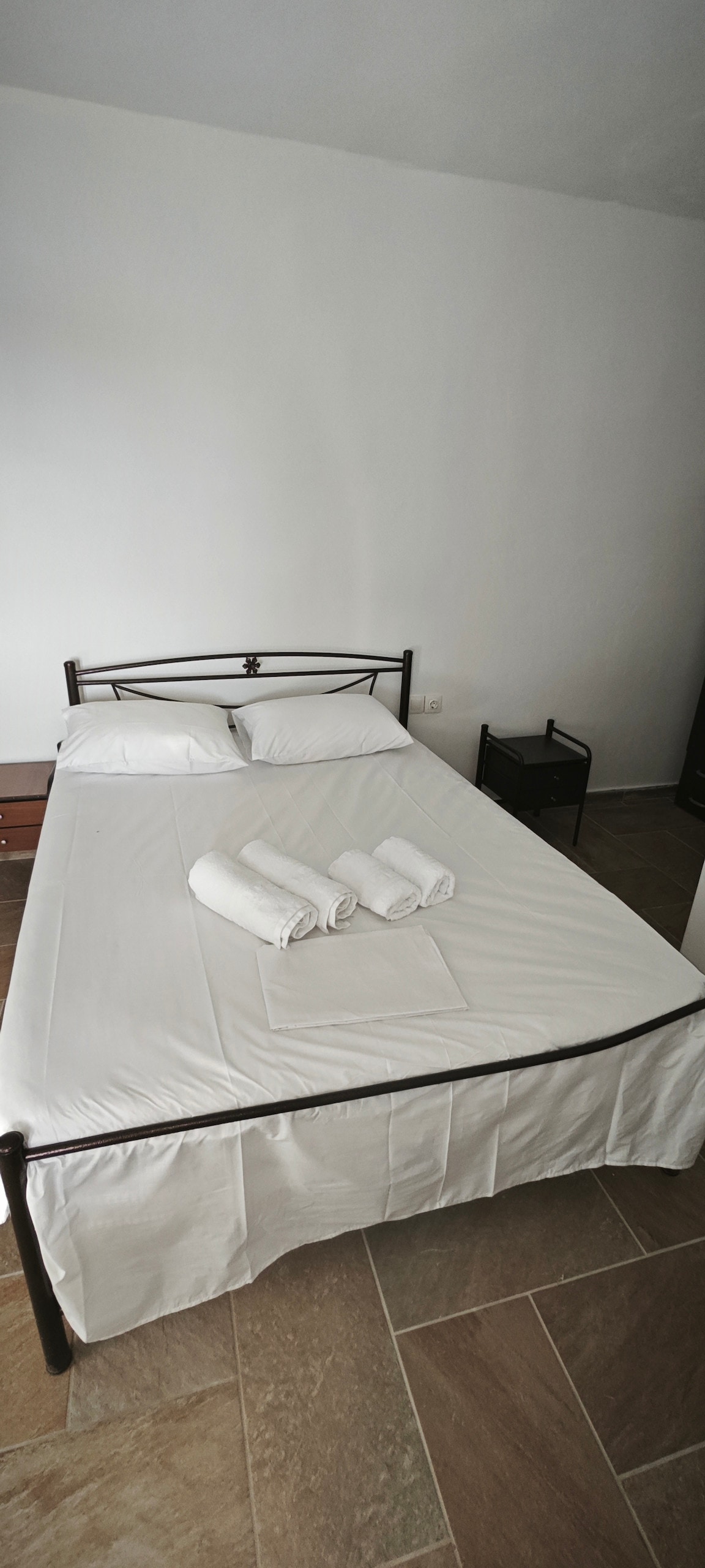 A spacious sleeping area features a metal frame bed dressed in crisp white linens. Two neatly arranged rolled towels sit at the foot of the bed. A small bedside table stands beside the bed, and the room is accented by light-colored tiled flooring.