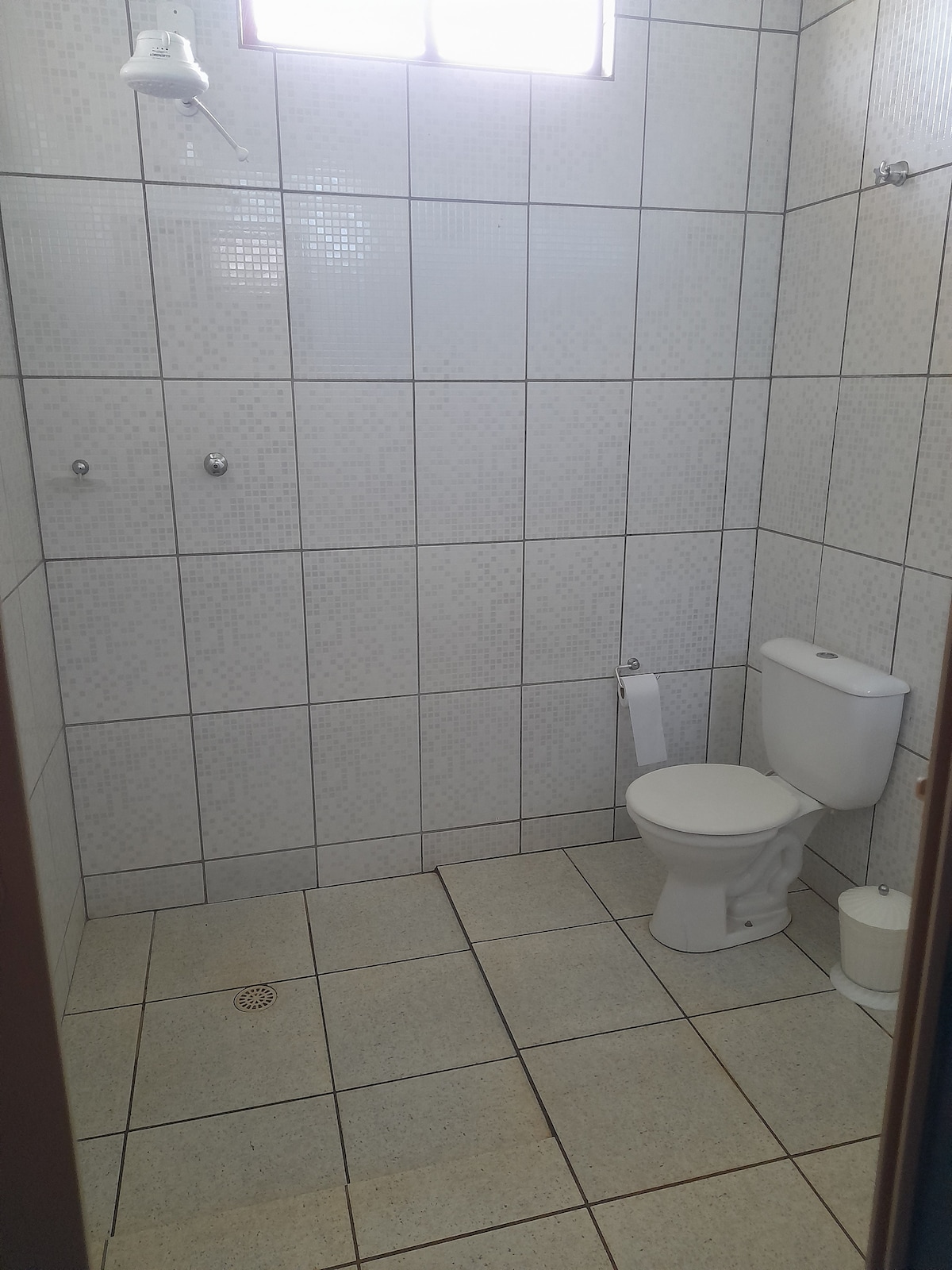 A tiled bathroom features neutral-colored tiles on the walls and floor. A white toilet is placed against one wall, while a shower area is equipped with a showerhead overhead. A small waste bin is located nearby, and natural light enters through a window.