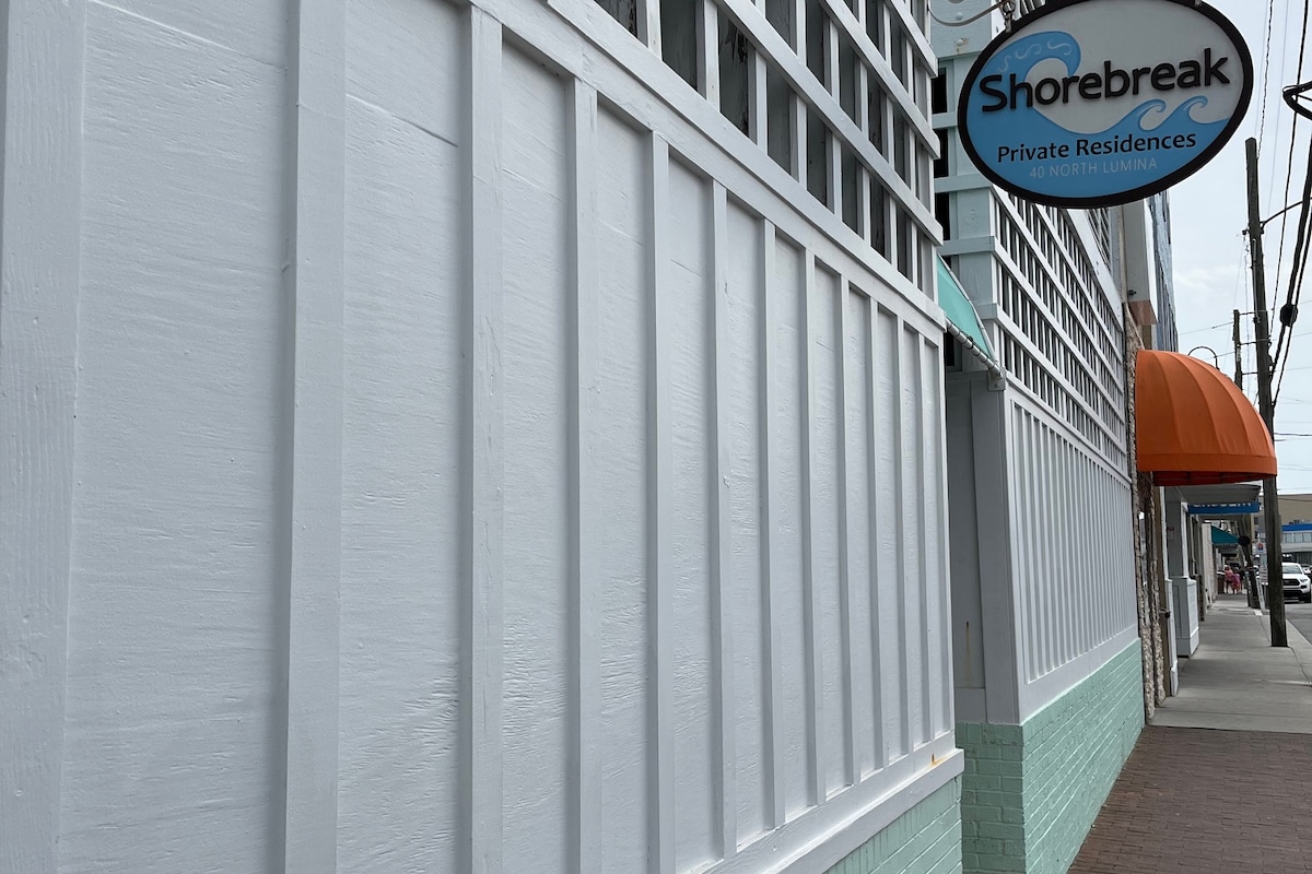 The exterior of a building named Shorebreak is displayed, featuring a white wooden wall with textured panels and a light turquoise base. An awning in orange can be seen above, along with a sign indicating private residences, set against a cloudy sky.