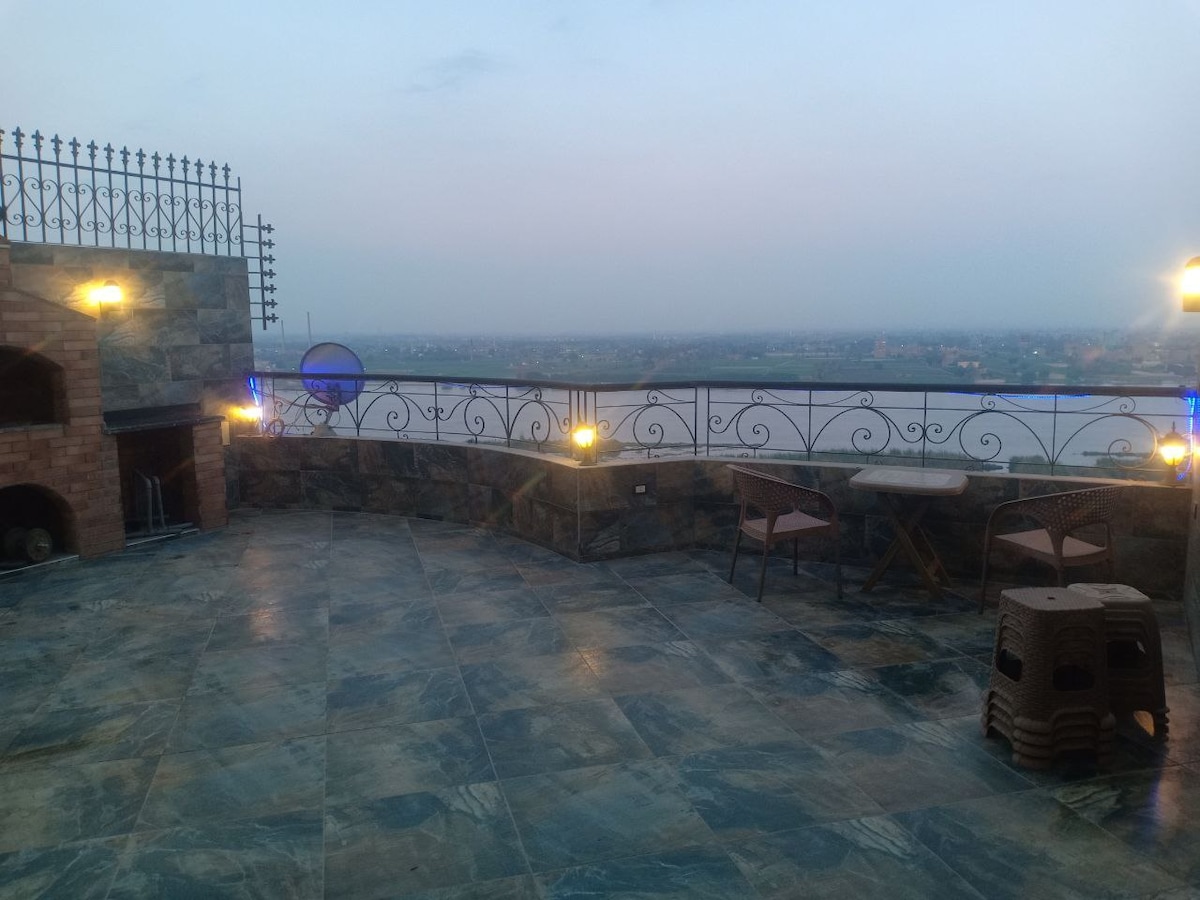 A spacious terrace is presented, adorned with illuminated railings and a scenic view of the Nile. Two wooden chairs and a small table are positioned on the tiled flooring, while decorative stone elements enhance the outdoor space.
