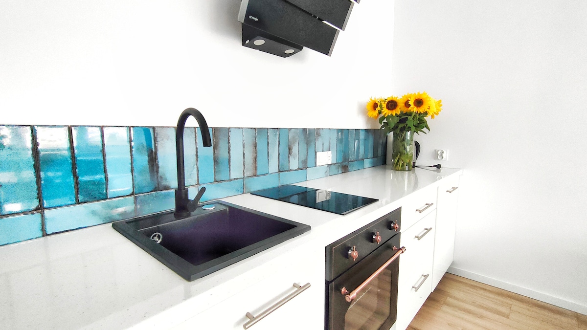 The kitchen features sleek white cabinetry and a modern countertop. A black sink and stovetop are positioned against a striking blue tile backsplash. A vase of sunflowers adds a touch of color, enhancing the space's bright and airy feel.