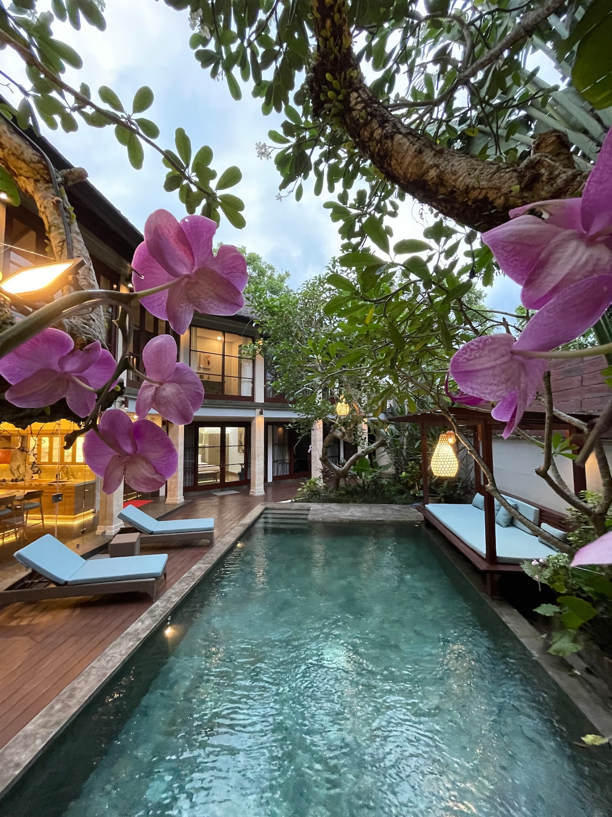 A serene pool area is framed by lush greenery and vibrant purple orchids. Sun loungers are positioned beside the sparkling water, reflecting the surrounding trees. A softly lit lantern hangs overhead, complementing the soothing ambiance of the outdoor space.