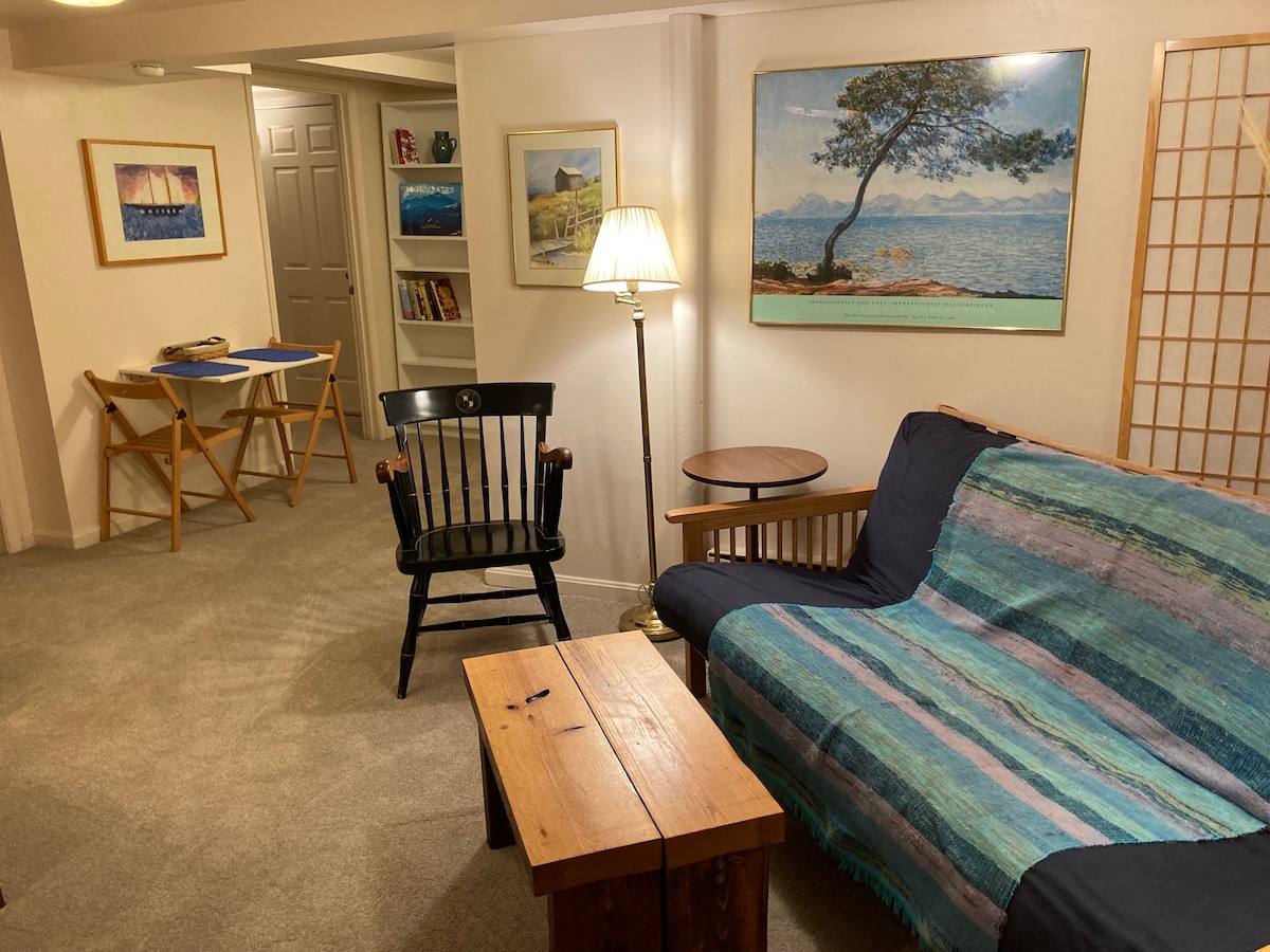 A comfortable seating area is highlighted by a wooden coffee table and a black rocking chair. Soft natural light is provided by a standing lamp, and serene artwork of landscapes adorns the walls. The cozy space is arranged for relaxation and leisure.
