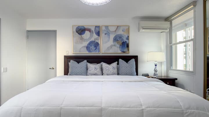 Sink into a luxurious king-size bed dressed in crisp white linens, plush pillows, and elegant blue accents, complemented by a rich wood headboard, modern abstract artwork, and stylish bedside lighting for a relaxing, upscale retreat.
