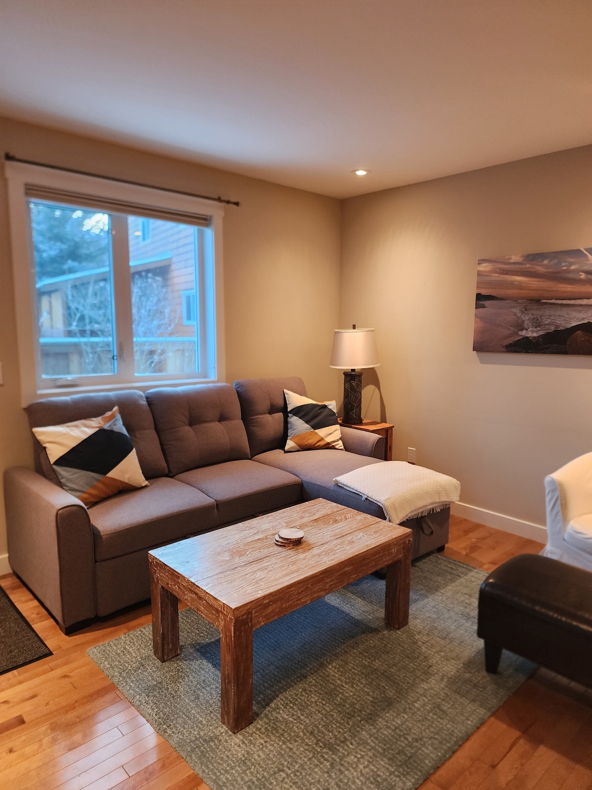 The cozy living space features a modern couch adorned with patterned cushions, complemented by a wooden coffee table and a textured area rug. Natural light flows in through a nearby window, illuminating the neutral-toned walls and enhancing the relaxed ambiance.