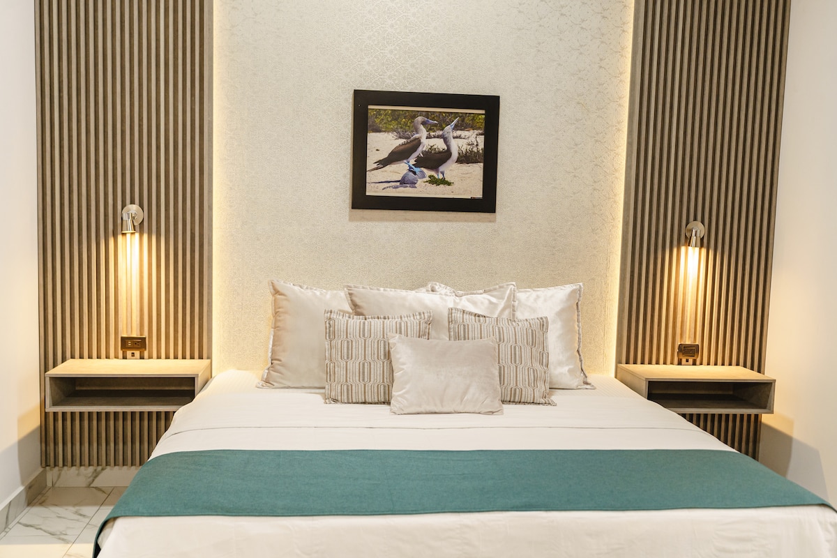 A spacious bedroom features a large bed with a light-colored duvet and a turquoise throw blanket. Soft lighting emanates from wall-mounted lamps beside the bed. A framed photograph of a serene outdoor scene adorns the wall, adding a touch of nature to the modern design.
