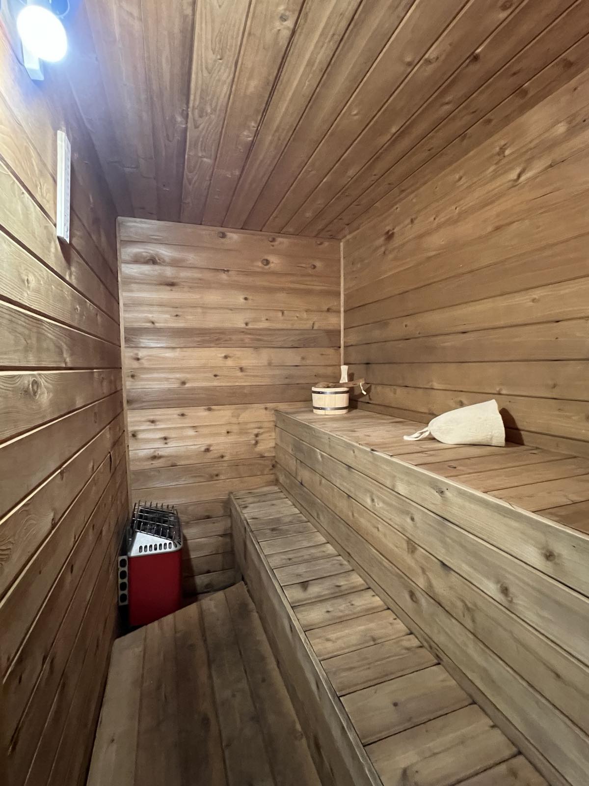 A private sauna is showcased with wood-paneled walls and benches, accommodating up to four individuals. A small wooden bucket and ladle are positioned on the bench, while a heater is placed discreetly in the corner, contributing to a relaxing environment.