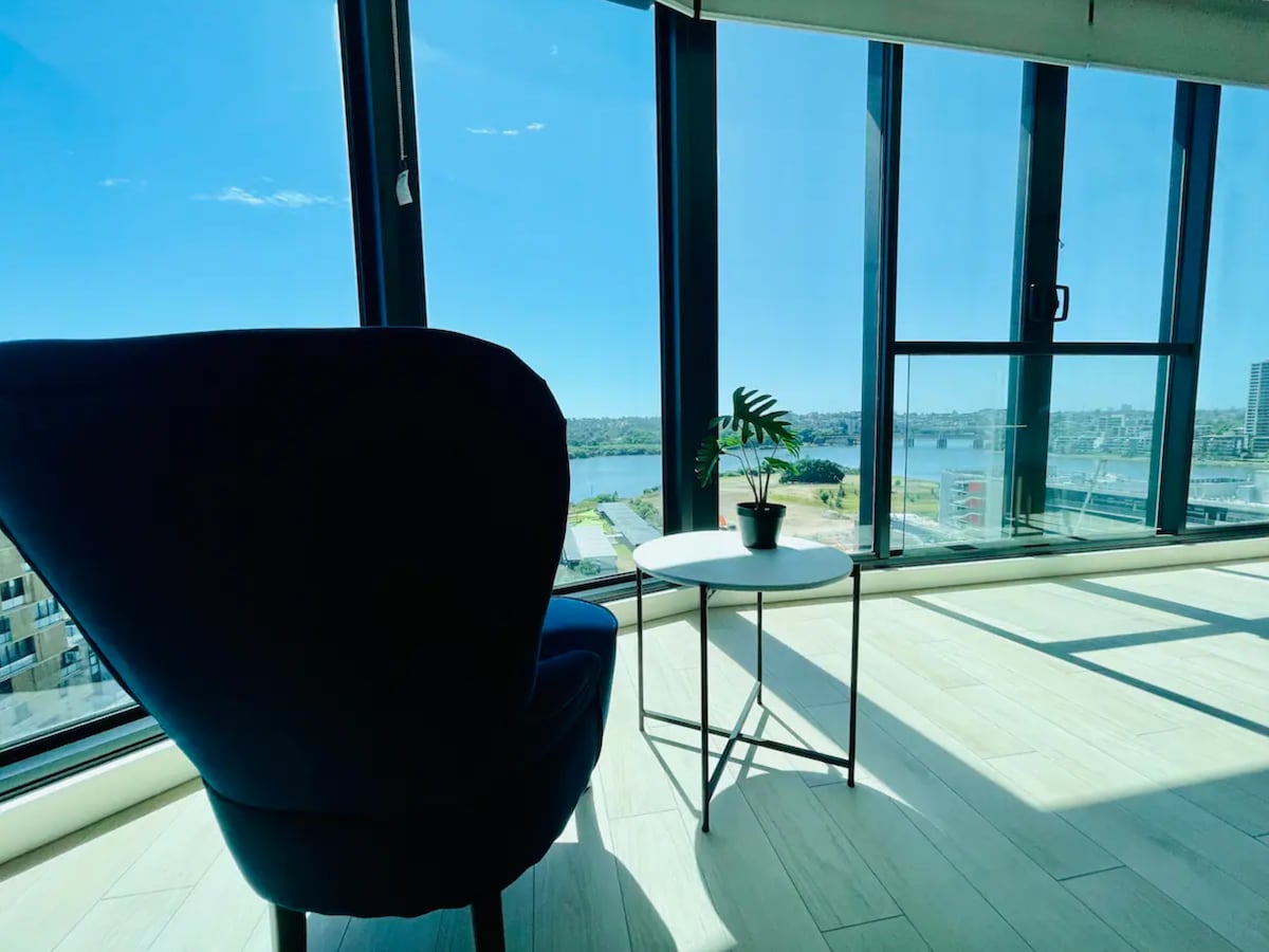 A modern armchair is positioned beside a small table, offering a comfortable spot to enjoy views through large floor-to-ceiling windows. Natural light illuminates the space, highlighting the minimalist decor and creating a welcoming atmosphere.