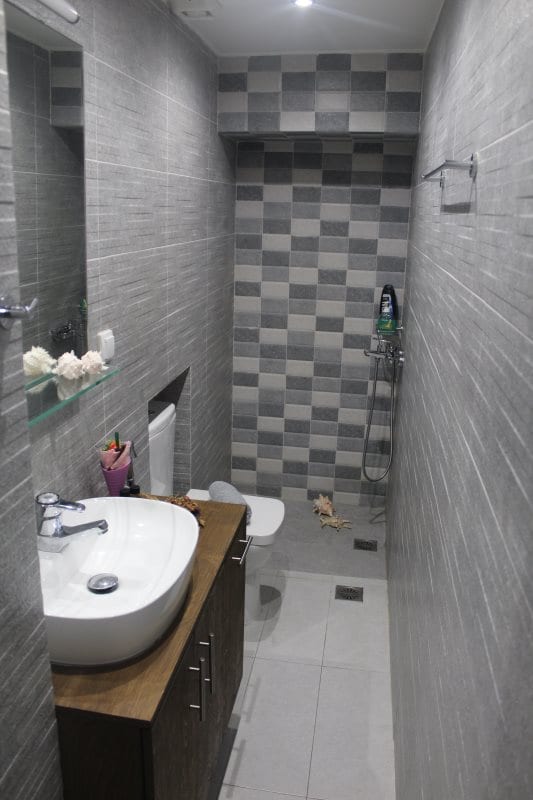A modern bathroom features grey tiled walls and a sleek design. A spacious sink is positioned above a dark wooden cabinet. The shower area has a handheld fixture, and personal care items are arranged neatly on a shelf. Floor tiles are presented in a light hue.