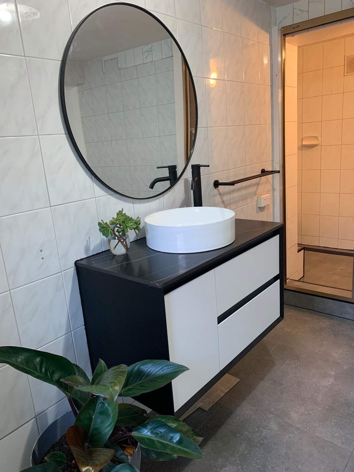 A modern bathroom setup is presented with a large round mirror above a sleek black and white vanity. A white vessel sink sits on the countertop. Natural light can be seen illuminating the shower area through a door behind. A small green plant adds a touch of nature.
