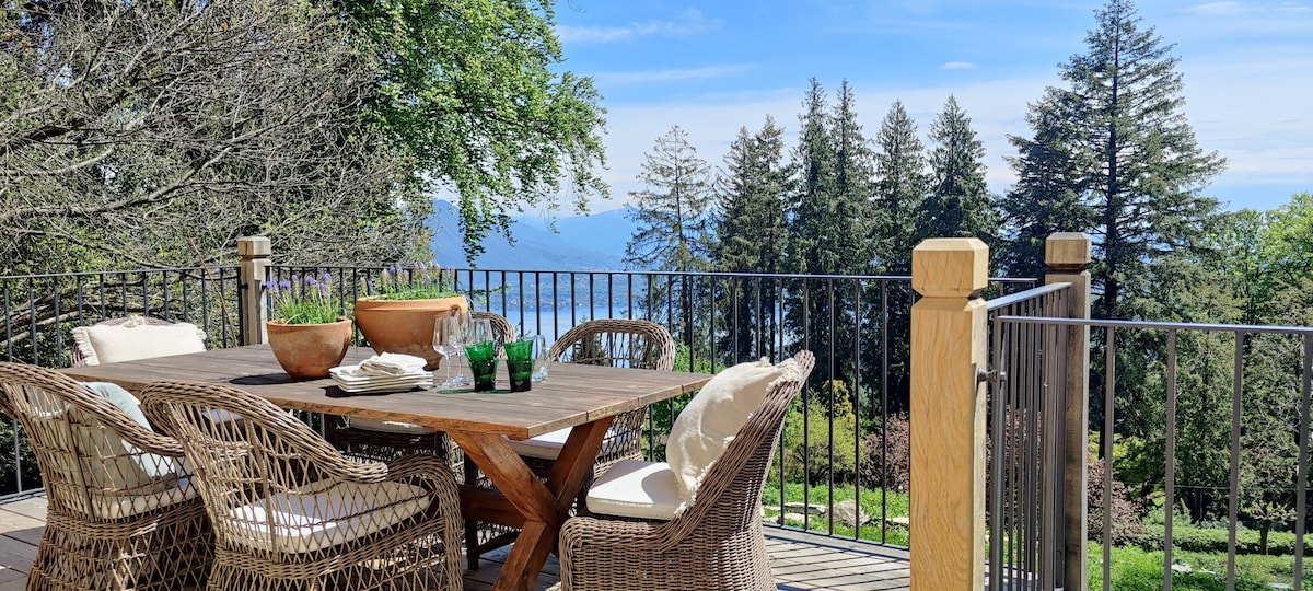 An outdoor dining area features a wooden table surrounded by woven chairs, with cushions for comfort. Beyond the railing, scenic views of Lake Maggiore and tall trees create a natural backdrop, inviting relaxation and connection with the surrounding landscape.