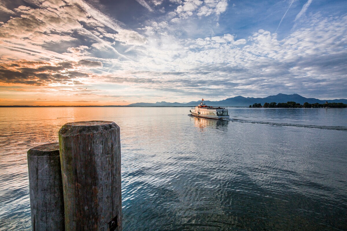 A tranquil scene shows a boat gliding over the calm waters of the Chiemsee at sunset. The sky is filled with soft clouds and warm hues, reflecting off the water. Wooden posts are visible in the foreground, anchored along the lakeshore.