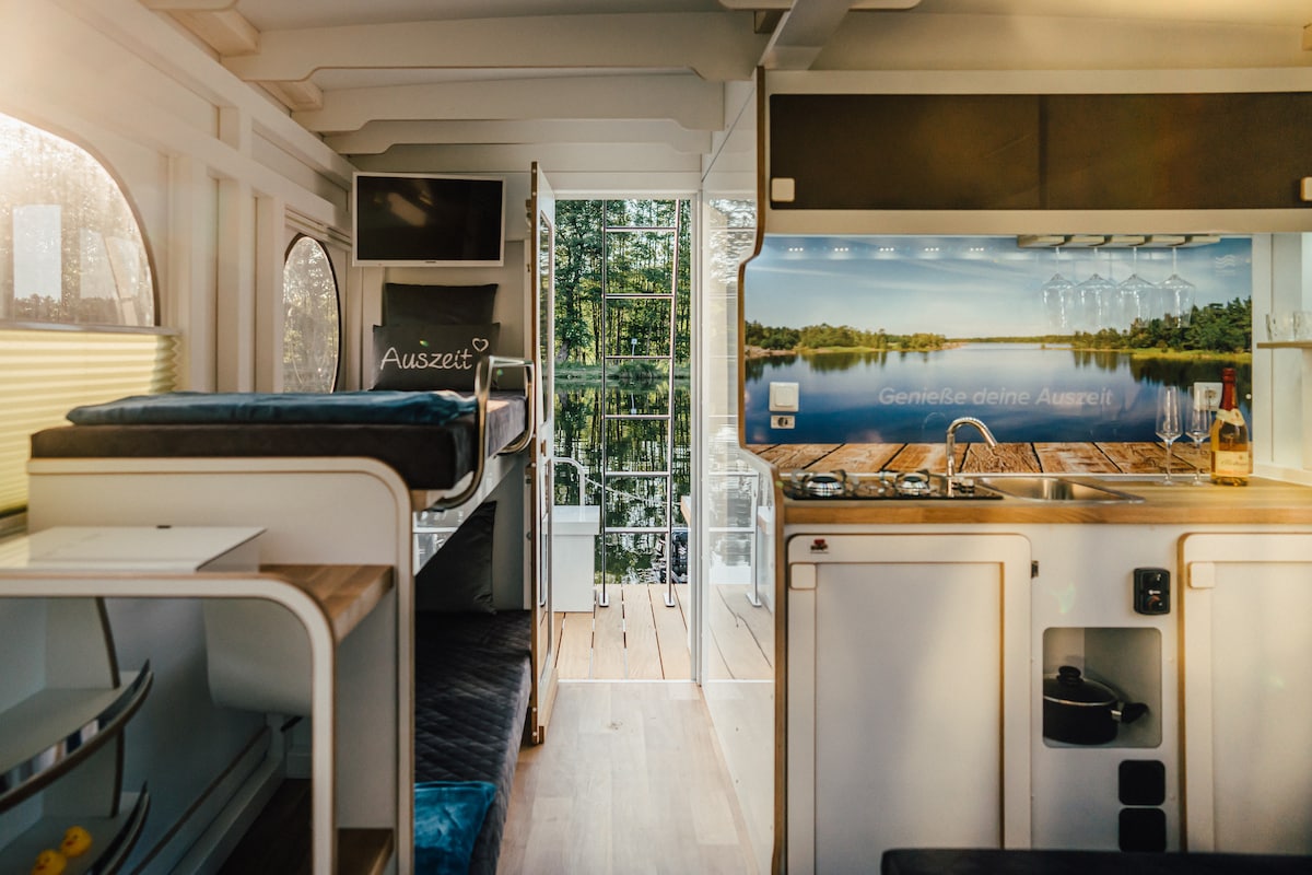 The interior of the boat features a bright and airy layout with wooden flooring and large windows. A compact kitchen area is equipped with modern appliances, while a spacious seating area offers flexibility for relaxation or dining. Natural light enhances the welcoming atmosphere.