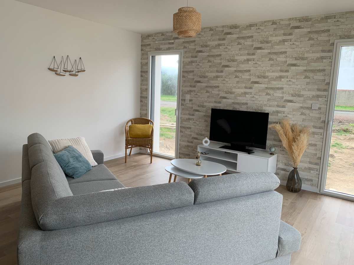A living area features a spacious gray sectional sofa with a decorative throw pillow. A light-colored coffee table is positioned in front, while a flat-screen television is placed on a modern console. Natural light enters through large windows, showcasing a simple and calm environment.