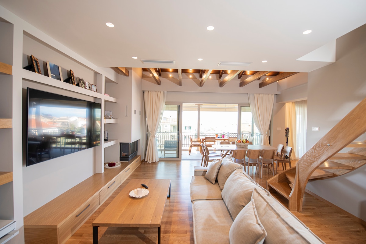 An open-plan living area is highlighted by warm wooden floors and beamed ceilings. A spacious seating arrangement faces a wall-mounted television, while natural light floods in through large sliding doors that lead to a balcony. A dining table is positioned in the background.
