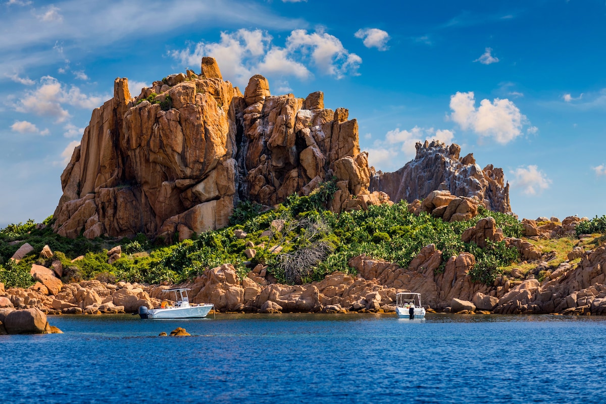 A rocky coastal formation rises from the clear blue water, featuring rugged cliffs and lush greenery. Two boats can be seen moored nearby, with the sky displaying scattered clouds and vibrant hues, creating a serene nautical scene.
