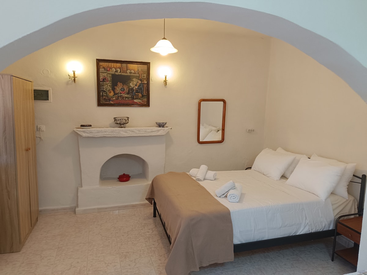 A spacious sleeping area features a bed covered with crisp linens and soft pillows. A rustic fireplace is positioned against a white wall, complemented by a simple wooden cabinet and a mirror. Warm lighting creates a cozy ambiance, enhancing the room's relaxation potential.