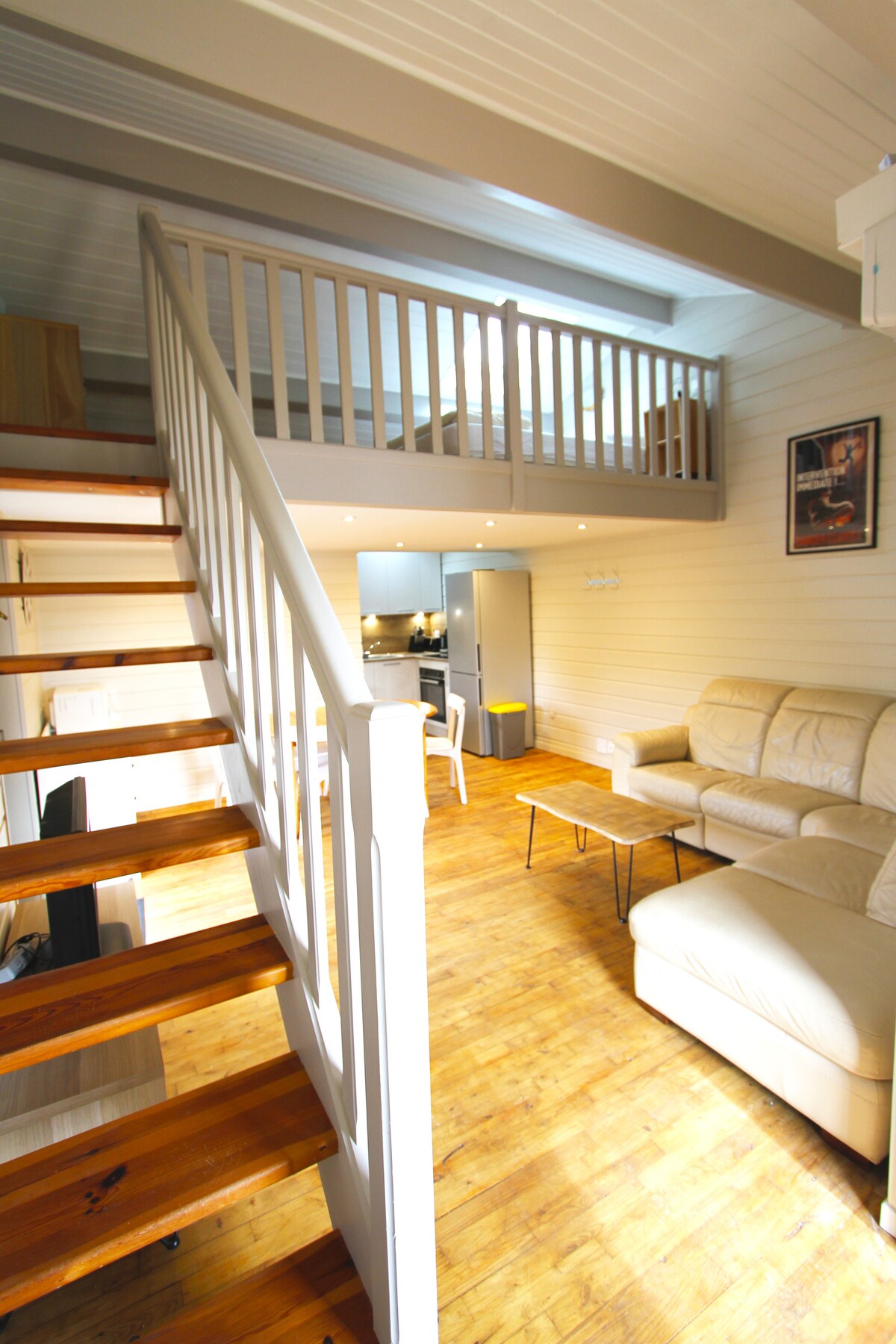 An open-plan interior showcases light wooden flooring and white wooden paneling. A comfortable beige sofa is positioned near a small coffee table. A staircase leads to a mezzanine level, while the kitchen area is visible in the background, illuminated by soft, recessed lighting.