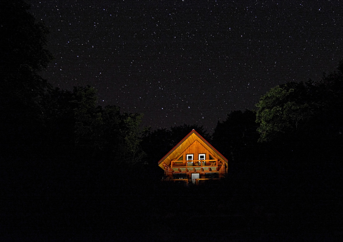 A secluded cabin is illuminated under a clear night sky filled with stars. The wooden structure features a steeply pitched roof and windows, with warm light glowing from within. Surrounded by dense trees, the cabin presents a peaceful retreat in nature.