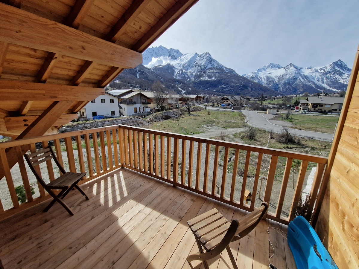 A wooden terrace is showcased, featuring two folding chairs positioned against the railing. A scenic mountain landscape is visible in the background, with a clear blue sky and snow-capped peaks complementing the view.