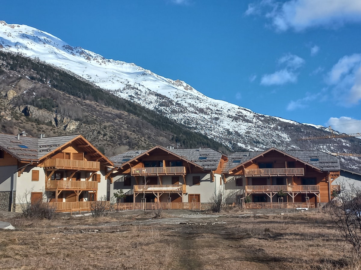 The image displays a serene mountain landscape featuring several wooden-residence structures, each with balconies. Snow-capped mountains serve as a backdrop, and the ground is covered with grass and sparse vegetation, indicating a peaceful setting. The sky is clear with scattered clouds.