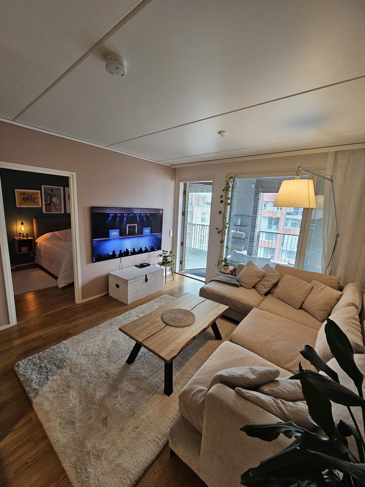 A spacious living area is seen, featuring a light-colored sectional sofa and a coffee table placed on a soft area rug. A large television is mounted on the wall, while natural light filters through the glass doors leading to a balcony.