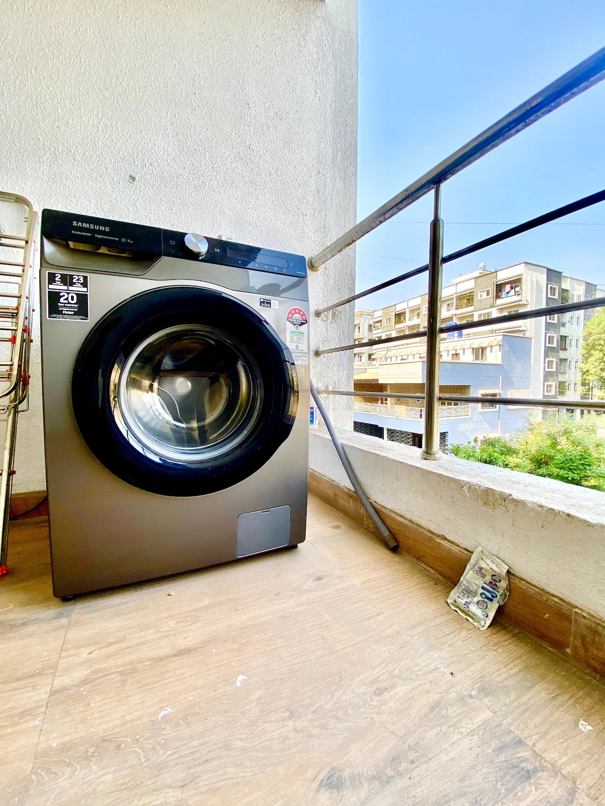 A fully automatic washing machine is positioned on the private balcony, showcasing a modern design and sleek finish. The background features a view of neighboring buildings bathed in natural light, while greenery peeks through, enhancing the outdoor space.